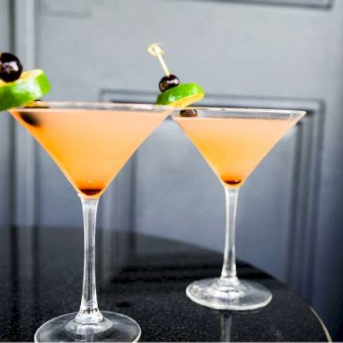 Two cocktails in martini glasses garnished with lime slices and cherries, set on a dark table.