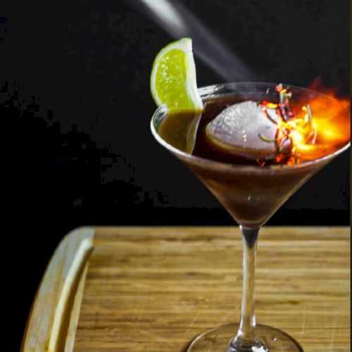 A cocktail in a martini glass on fire, decorated with a lime, and a torch being used above it on a wooden surface.