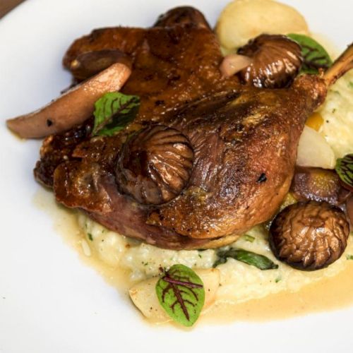 It’s a dish with roasted meat, mushrooms, pears, and greens on a bed of creamy mashed potatoes on a white plate.