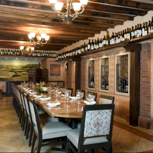 A rustic dining room with a long wooden table, elegantly set. Wine bottles line the ceiling, with paintings and warm lighting enhancing the ambiance.