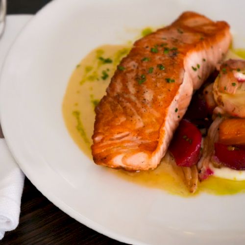A plate with grilled salmon, roasted vegetables, and a drizzle of sauce, alongside a fork and knife on a white napkin.