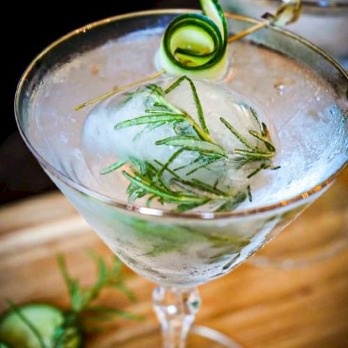 A martini glass filled with a clear cocktail, garnished with a rosemary sprig and a cucumber ribbon.