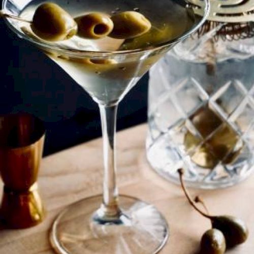 A martini with olives on a wooden surface alongside a cocktail shaker, measuring jigger, and yellow flowers.