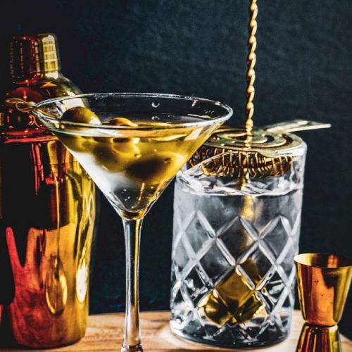 A martini with olives is on a wooden board, surrounded by a cocktail shaker, mixing glass with a spoon, and a jigger.