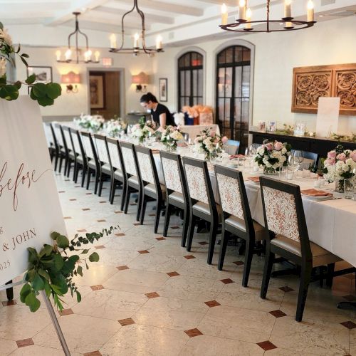 The image shows an elegantly set dining room with a sign for an event celebrating Jacqueline and John, adorned with floral arrangements.