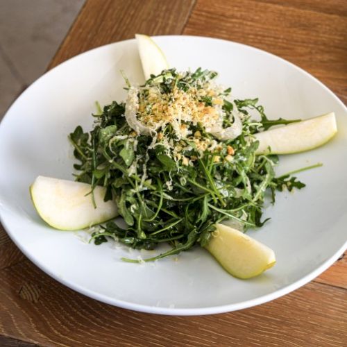 A salad with arugula, pear slices, grated cheese, and breadcrumbs is presented on a white plate on a wooden table.