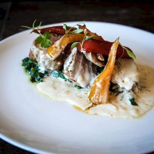 A gourmet dish featuring fish with creamy sauce, sautéed greens, and roasted vegetables, garnished with herbs on a white plate.