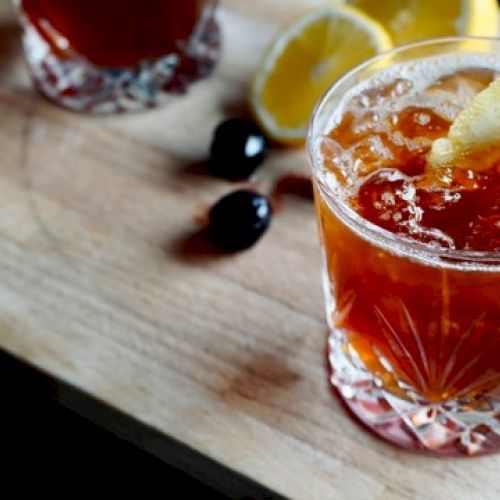 A cocktail in a glass with ice, garnished with lemon and cherry on a skewer, lemons and another glass in the background.
