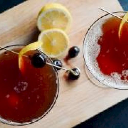 Two cocktails with cherries and lemon twists on a wooden board, accompanied by lemon slices and extra cherries.