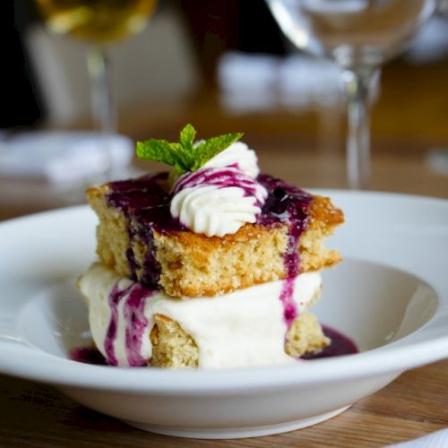 A layered dessert with cake, ice cream, berry sauce, whipped cream, and a mint garnish is on a plate, with wine glasses in the background.