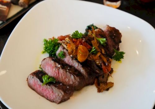 A plate of sliced steak with garnish, accompanied by bottles of whiskey, glasses, and cigars on the table.