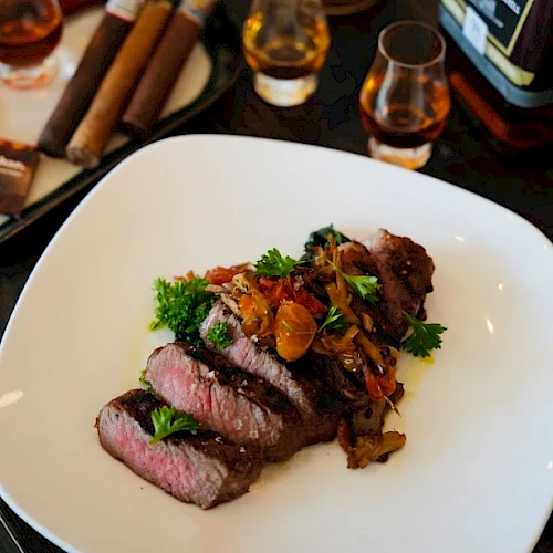A plate of sliced steak with garnish, accompanied by bottles of whiskey, glasses, and cigars on the table.