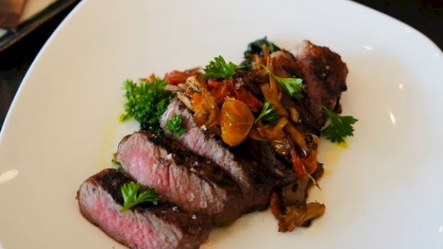 A plate of sliced steak with garnish, accompanied by bottles of whiskey, glasses, and cigars on the table.