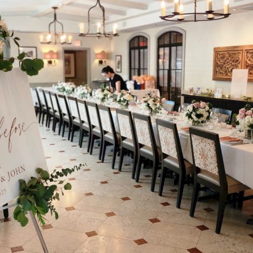 The image shows an elegantly set dining table in a decorated room, with a floral sign and stylish chairs, ready for an event.