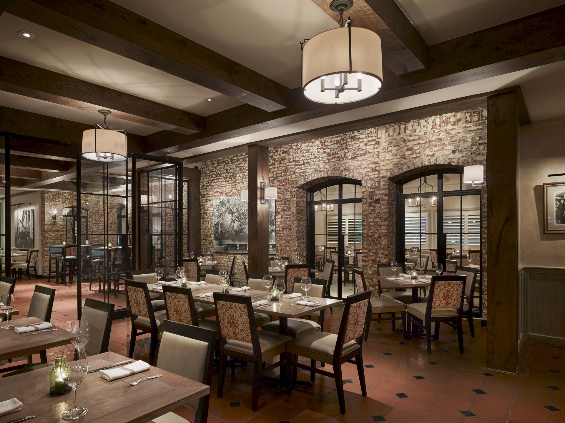 A cozy restaurant interior with wooden beams, brick walls, elegant lighting, and neatly arranged tables and chairs.