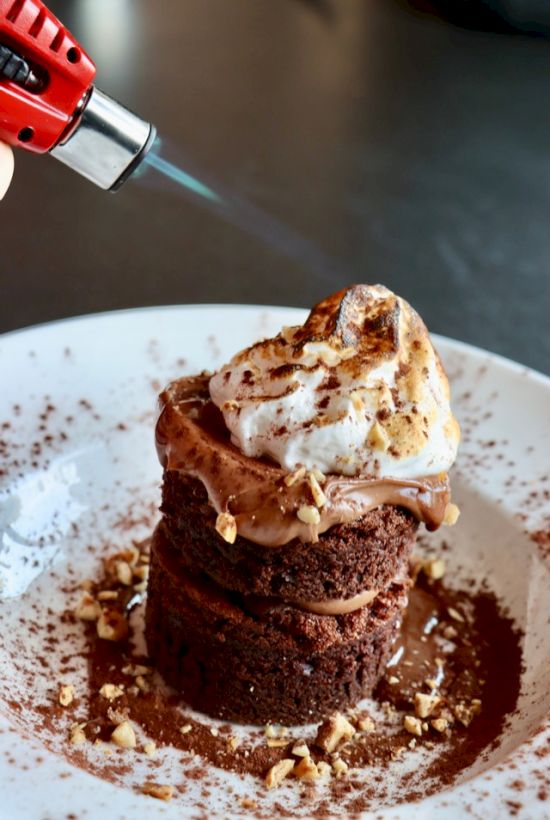 A chocolate dessert being torched, topped with whipped cream and nuts on a white plate, sprinkled with cocoa powder.