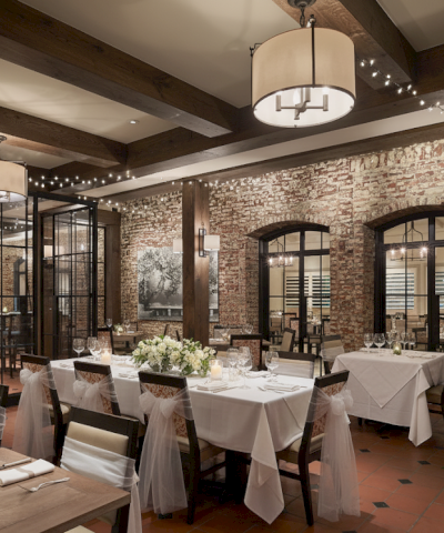 A warmly lit restaurant with brick walls, set for a wedding with white tablecloths, floral arrangements, and hanging string lights.