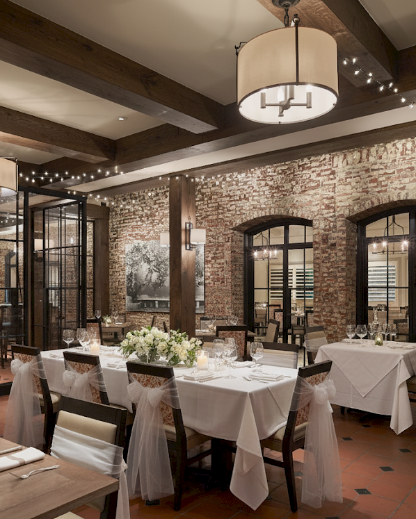 A warmly lit restaurant with brick walls, set for a wedding with white tablecloths, floral arrangements, and hanging string lights.