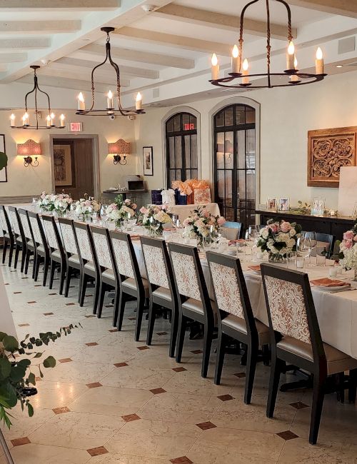 A long, elegant wedding reception setup with a white-clothed head table and floral centerpieces, chandeliers, and a welcome sign at the entrance.