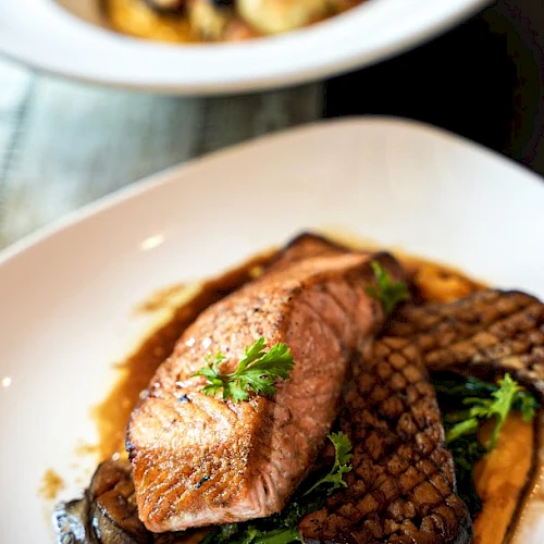 A plated dish with grilled salmon, greens, and sauce, with another dish blurred in the background, garnished with herbs.
