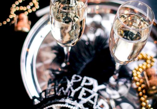 Two champagne flutes on a shiny tray with festive beads and a &ldquo;Happy New Year&rdquo; headband, suggesting a celebratory party vibe.