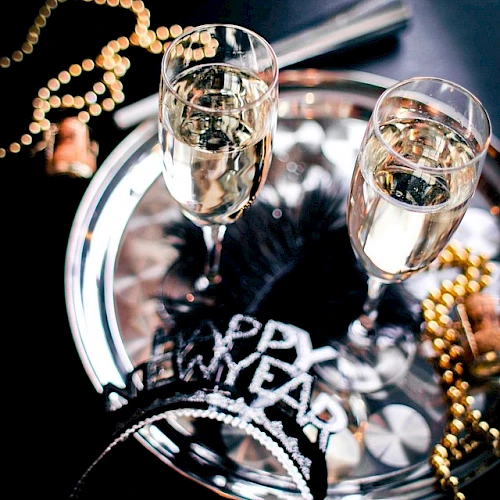Two champagne flutes on a shiny tray with festive beads and a &ldquo;Happy New Year&rdquo; headband, suggesting a celebratory party vibe.
