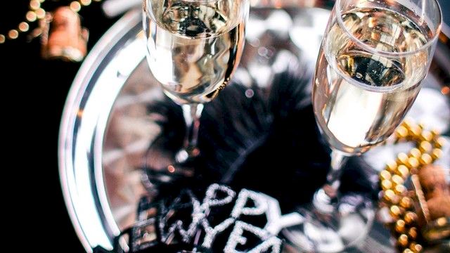Two champagne flutes on a shiny tray with festive beads and a “Happy New Year” headband, suggesting a celebratory party vibe.