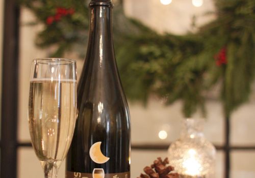 A bottle of dark beer or sparkling wine, a glass, a pinecone, a lit candle, and some greenery in the background, set on a wooden table.