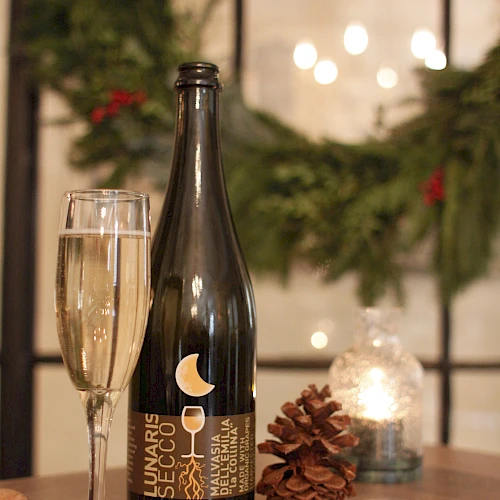 A bottle of dark beer or sparkling wine, a glass, a pinecone, a lit candle, and some greenery in the background, set on a wooden table.