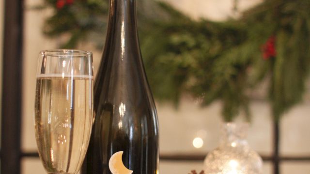 A bottle of dark beer or sparkling wine, a glass, a pinecone, a lit candle, and some greenery in the background, set on a wooden table.