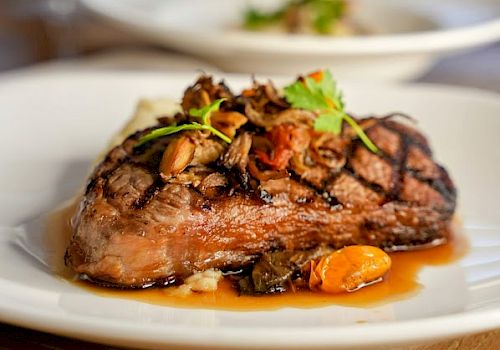 A plated steak topped with mushrooms and greens, served with a savory glaze on a white dish, wine glass in the blurred background.