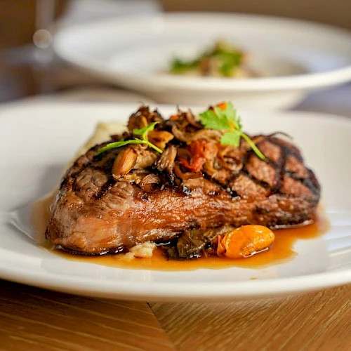 A plated steak topped with mushrooms and greens, served with a savory glaze on a white dish, wine glass in the blurred background.