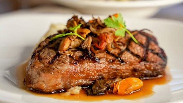 A plated steak topped with mushrooms and greens, served with a savory glaze on a white dish, wine glass in the blurred background.