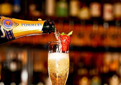 Champagne being poured into a flute garnished with a strawberry, set against a bar backdrop with another strawberry on the counter. The sentence ends here.