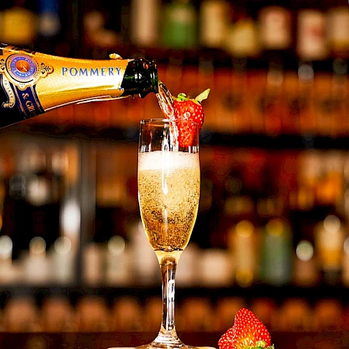 Champagne being poured into a flute garnished with a strawberry, set against a bar backdrop with another strawberry on the counter. The sentence ends here.