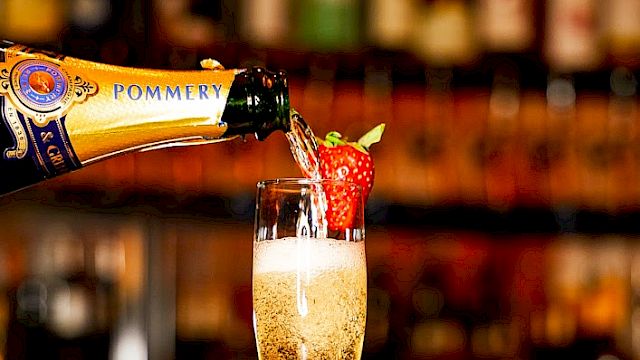 Champagne being poured into a flute garnished with a strawberry, set against a bar backdrop with another strawberry on the counter. The sentence ends here.