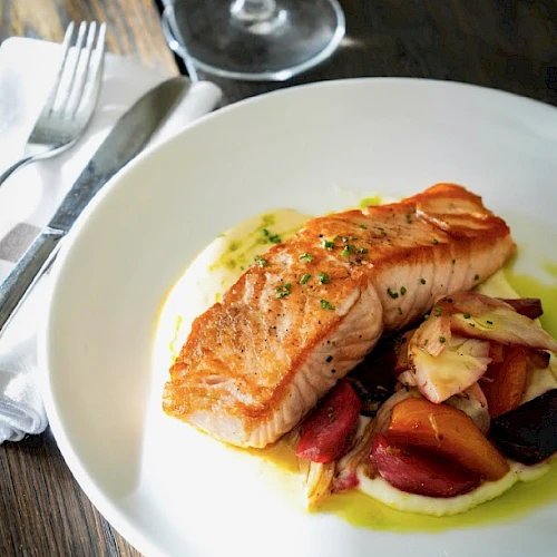 A plated salmon fillet with a light sear sits on creamy mashed potatoes, surrounded by roasted vegetables, and a sprig of herbs on top.