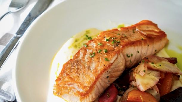 A plated salmon fillet with a light sear sits on creamy mashed potatoes, surrounded by roasted vegetables, and a sprig of herbs on top.