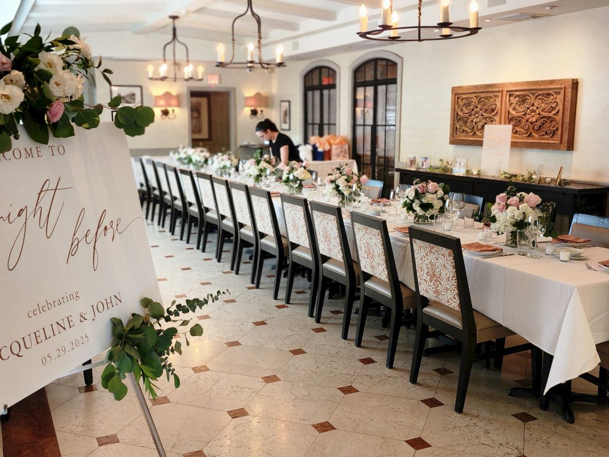A long dining table setup for a wedding reception with a sign reading &ldquo;the night before&rdquo; and floral centerpieces along the table.