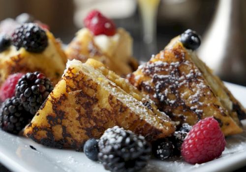 Plate of French toast with berries, dusted sugar, and a splash of maple; a glass of orange juice in the background, delicious breakfast vibes.