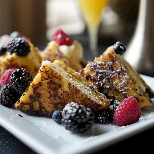 Plate of French toast with berries, dusted sugar, and a splash of maple; a glass of orange juice in the background, delicious breakfast vibes.