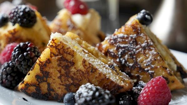 Plate of French toast with berries, dusted sugar, and a splash of maple; a glass of orange juice in the background, delicious breakfast vibes.