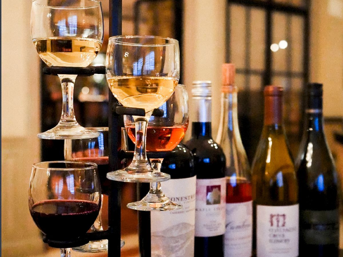 A tiered wine flight with three glasses of white and red wine, plus several bottles behind, arranged on a dark wooden table in a cozy setting.