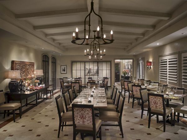 A styled formal dining room with a chandelier, candlelit tables, patterned chairs, and large windows and shutters.