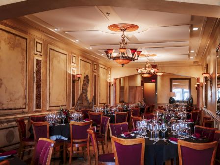 Elegant dining room with ornate walls, chandeliers, and round tables set with glassware and red chairs, ready for a formal event.