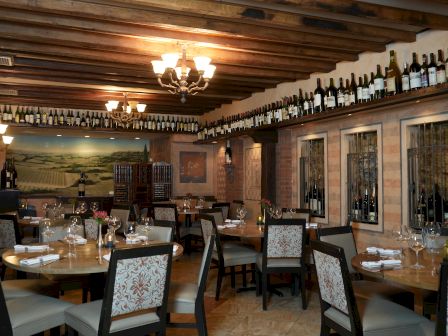 A cozy dining room with wooden beams, chandeliers, framed windows, and round tables set for dinner, bottles line the ceiling edge.