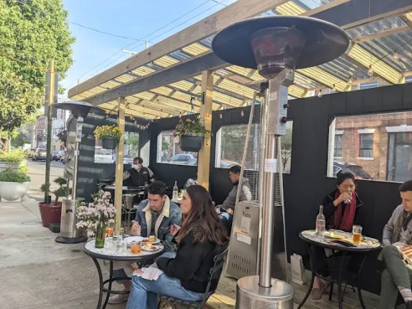 People dine at an outdoor café under patio heaters, with flowers in planters and several customers seated at tables.