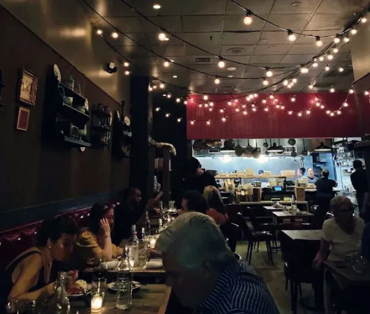 A cozy restaurant dining room with warm string lights, people seated at tables, and a small bar in the background.