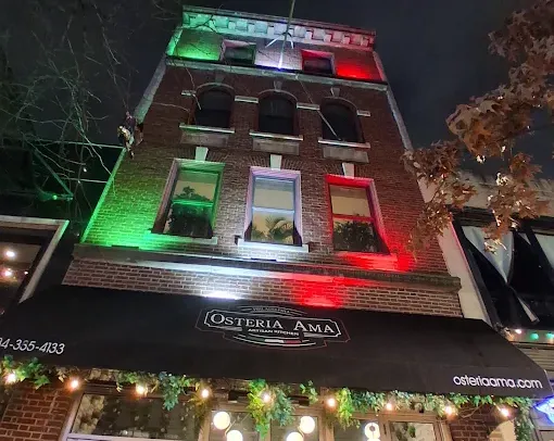 A nighttime view of a brick restaurant facade, glowing green, red, and blue lights, with the sign “Osteria AMA” above the entrance.