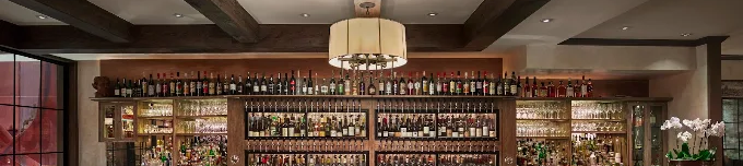 A cozy bar with a long wooden counter, many bottles on shelves, stools, warm lighting, and a hanging chandelier.
